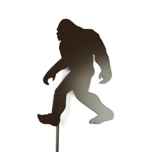 May include: A metal silhouette of a Bigfoot figure walking. The figure is dark brown with a gradient effect, mounted on a thin metal rod. The background is plain white.