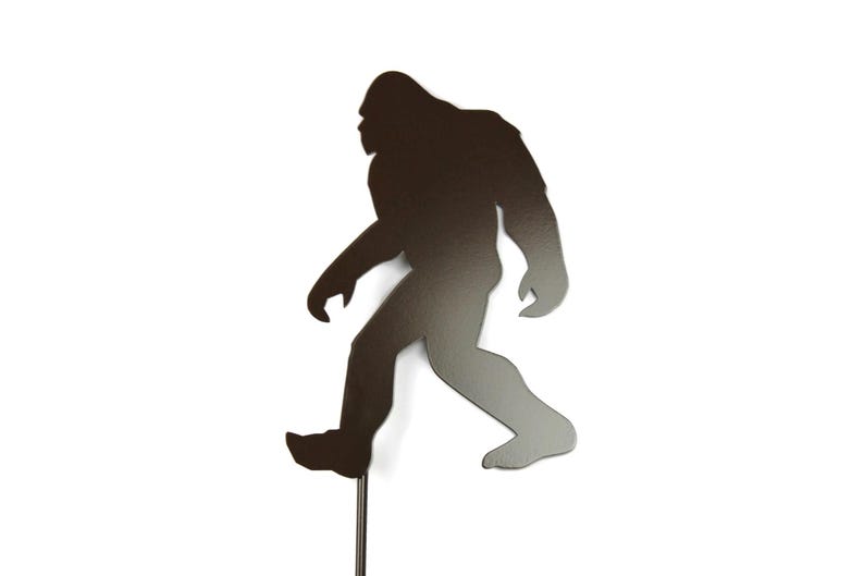 May include: A metal silhouette of a Bigfoot figure walking. The figure is dark brown with a gradient effect, mounted on a thin metal rod. The background is plain white.