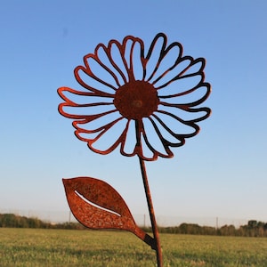 May include: A rustic metal daisy garden stake with a detailed cutout design. The flower is a rusty brown color, with a stem and leaf. The stake is placed in a grassy field with a blue sky background.