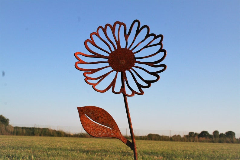 May include: A rustic metal daisy garden stake with a detailed cutout design. The flower is a rusty brown color, with a stem and leaf. The stake is placed in a grassy field with a blue sky background.