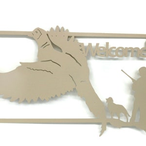May include: Tan metal wall art featuring a duck in flight, a hunter with a rifle, and a dog. The word "Welcome" is cut out above the hunter. The art is framed with a rectangular border.