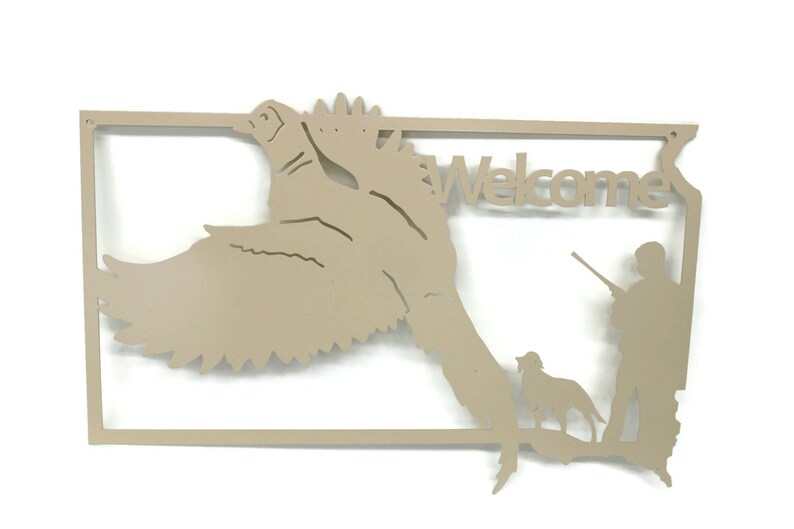 May include: Tan metal wall art featuring a duck in flight, a hunter with a rifle, and a dog. The word "Welcome" is cut out above the hunter. The art is framed with a rectangular border.