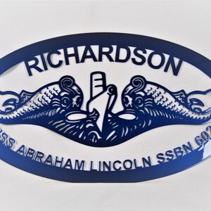 May include: A blue oval metal sign with the text "USS Abraham Lincoln SSBN 602" and "Richardson" in a white font. The sign features a stylized image of a submarine with two dolphins flanking it.