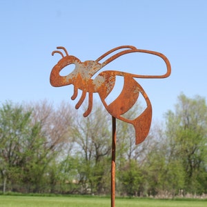 May include: A rusty metal bee silhouette garden stake. The bee is in flight with wings spread and has a striped body. The stake is brown and is set against a background of green trees and a blue sky.