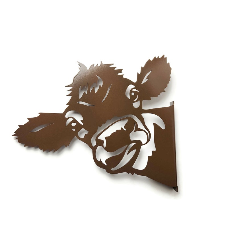 May include: A brown metal cow head wall decor with a playful expression. The cow's head is cut out of metal and has a brown finish.
