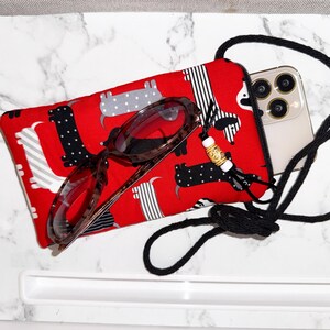 May include: A red fabric phone case with a black zipper, featuring a dachshund pattern in black, white, and gray. Tortoiseshell eyeglasses rest on the case, and a black cord is attached. A smartphone is visible inside the case.