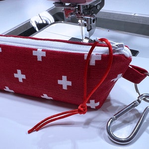 May include: A red zippered pouch with a white cross pattern and a red drawstring. The pouch has a silver carabiner clip attached to a silver ring.