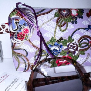 May include: A purple fabric pouch with a floral design and gold accents. The pouch has a purple drawstring and a silver clip. The pouch is sitting on a white surface with a pair of glasses.
