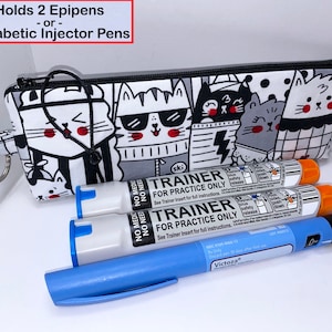 May include: A black and white zippered pouch with a cartoon cat design. The pouch is labeled "Holds 2 Epipens - or - 3 Diabetic Injector Pens". The pouch is shown with three medical injector pens, two with orange caps and one with a blue cap.