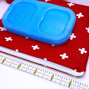 May include: Red zippered pouch with white plus signs printed on the fabric. A blue ice pack is on top of the pouch. A white ruler with yellow markings is in the foreground.