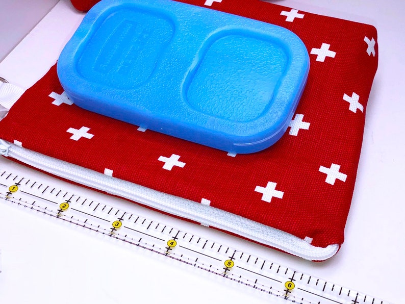 May include: Red zippered pouch with white plus signs printed on the fabric. A blue ice pack is on top of the pouch. A white ruler with yellow markings is in the foreground.