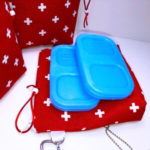 May include: Two blue rectangular ice packs with a divided compartment on top of a red fabric pouch with white plus signs. The pouch has a white drawstring and a silver chain attached.