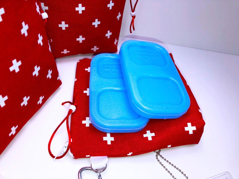 May include: Two blue rectangular ice packs with a divided compartment on top of a red fabric pouch with white plus signs. The pouch has a white drawstring and a silver chain attached.