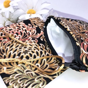May include: Two small, black fabric pouches with a floral pattern in gold, red, and green. One pouch is open, revealing a white interior. The pouches have a gold drawstring closure and a black zipper.