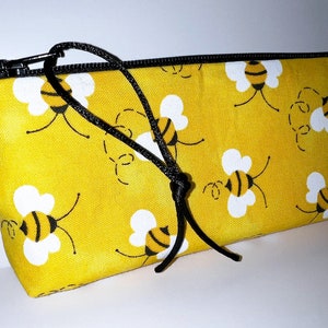 May include: Yellow pencil case with a zipper closure and a black drawstring. The case is decorated with a repeating pattern of cartoon bees.
