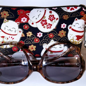 May include: A black fabric pouch with a Japanese lucky cat design and a red zipper pull. The pouch is lying on a white surface next to a pair of brown tortoise shell sunglasses.