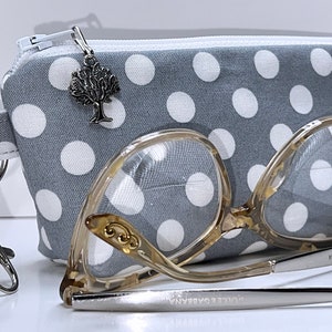 May include: A gray and white polka dot fabric eyeglasses case with a zipper closure and a silver tree charm. The case is lying on a white surface next to a pair of clear framed eyeglasses with a silver metal frame and the text "DOLCE & GABBANA" on the side.