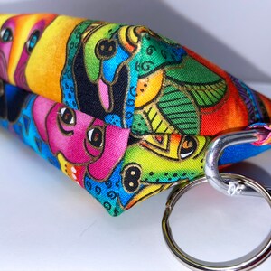 May include: A colorful fabric pouch with a whimsical design featuring cartoon faces and abstract patterns. The pouch has a silver metal key ring and a carabiner clip.