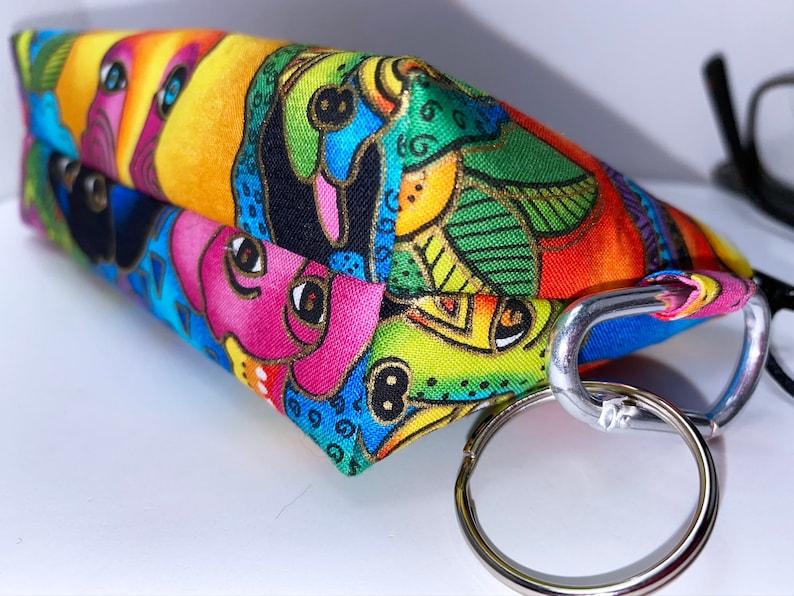 May include: A colorful fabric pouch with a whimsical design featuring cartoon faces and abstract patterns. The pouch has a silver metal key ring and a carabiner clip.