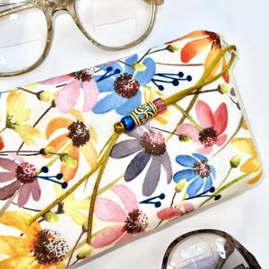 May include: A white floral patterned glasses case with a gold tassel and a blue and gold bead. The case is closed and lying on a white surface.