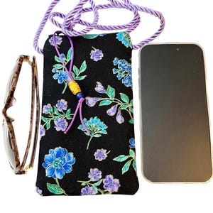 May include: A black floral print phone bag with a purple rope strap. The bag features blue and purple flowers with green leaves. Tortoiseshell eyeglasses and a smartphone are next to the bag.