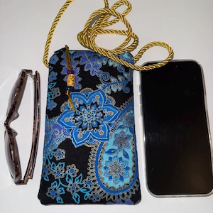May include: A black and blue floral patterned phone case with a gold rope strap. The case is next to a pair of brown sunglasses and a black smartphone. The case has a blue and gold floral design.