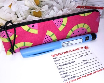 Pode incluir: Uma bolsa rosa com zíper com um design de estampa de melancia. A bolsa contém uma caneta de insulina azul com a etiqueta "Victoza". Também é possível ver um cartão de informações médicas de emergência.