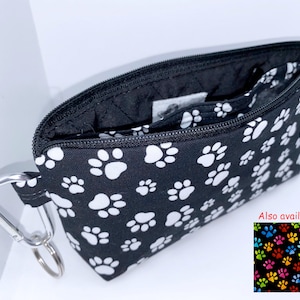 May include: Black zippered pouch with a white paw print pattern. The pouch has a silver carabiner clip and a zipper pull with a wooden bead. The pouch is open and the inside is lined with black fabric. Also available: Multicolored paw print pattern.