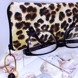 Pode incluir: Um estojo para &oacute;culos de estampa de leopardo com fecho de correr e um par de &oacute;culos pretos com lentes transparentes. O estojo e os &oacute;culos est&atilde;o sobre uma superf&iacute;cie branca com conchas.