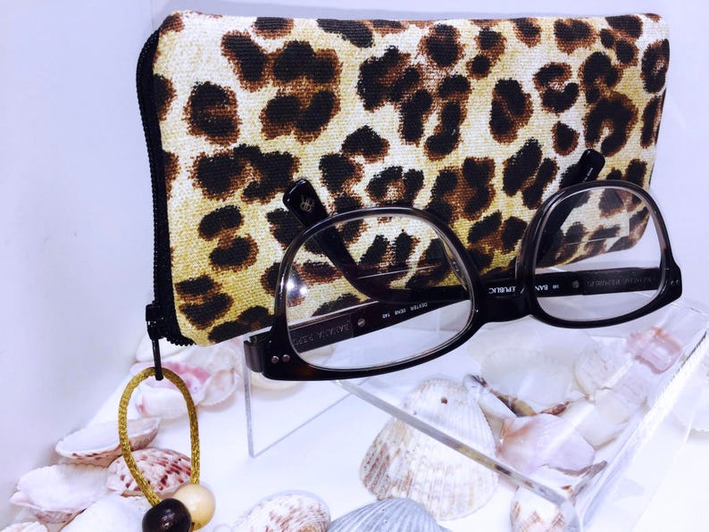 Pode incluir: Um estojo para &oacute;culos de estampa de leopardo com fecho de correr e um par de &oacute;culos pretos com lentes transparentes. O estojo e os &oacute;culos est&atilde;o sobre uma superf&iacute;cie branca com conchas.