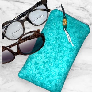 May include: A teal floral print glasses case with a black zipper and a beaded tassel next to two pairs of eyeglasses. One pair has a black and tortoiseshell frame, and the other has a brown tortoiseshell frame.