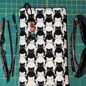 May include: Black and white cat print fabric eyeglasses case with a black cord and colorful beads. The case is lying on a green cutting mat with two pairs of eyeglasses.