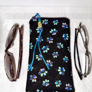 May include: A black eyeglass case with a blue paw print pattern, a blue tassel, and two pairs of eyeglasses. One pair has a tortoise shell pattern, and the other has silver frames.