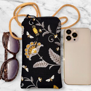 May include: Black phone crossbody bag with a floral pattern in yellow, white, and gray. The bag has a long tan cord strap and a zipper closure. A pair of sunglasses and a gold phone are next to the bag.