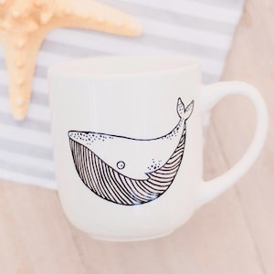 Op de afbeelding: Witte keramische mok met een zwarte lijntekening van een walvis op de voorkant. De walvis kijkt naar links en heeft een klein oog.