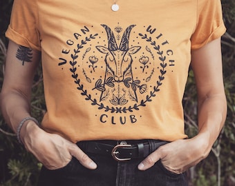 Op de afbeelding: Een mosterdgeel T-shirt met een zwarte afbeelding van een geitenkop met een krans van bloemen eromheen. De tekst "Vegan Witch Club" is rond de krans gedrukt.