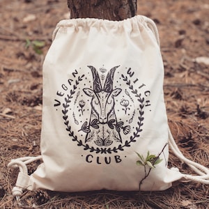 Op de afbeelding: Een witte trekkoordtas met een zwart-witte afbeelding van een geitenkop met bloemmotieven. De tekst "Vegan Witch Club" is rond de afbeelding gedrukt.