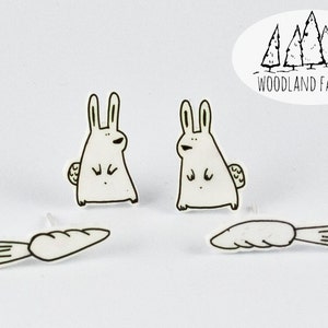 May include: A pair of white bunny rabbit earrings with a carrot charm. The bunny rabbits are drawn in a simple, cartoon style. The earrings are made by Woodland Factory.
