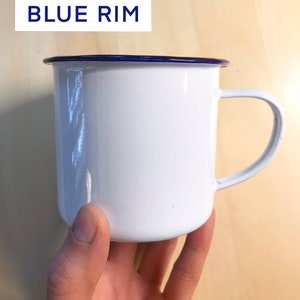 May include: White enamel mug with a blue rim and a handle.  The mug is being held by a hand.
