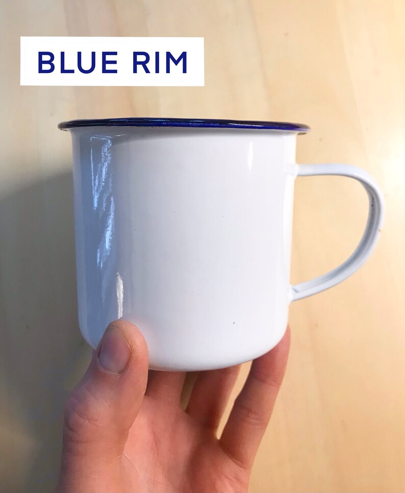 May include: White enamel mug with a blue rim and a handle.  The mug is being held by a hand.