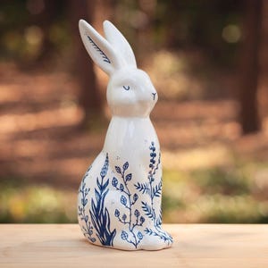Może przedstawiać: Biała ceramiczna figurka królika z błyszczącym wykończeniem i misternymi niebieskimi ilustracjami botanicznymi liści, łodyg i jagód na ciele. Królik ma długie uszy, zamknięte oczy i mały nos, wszystko wykonane niebieskim tuszem.