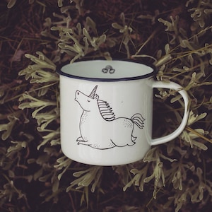 Könnte beinhalten: Weiße Email Tasse mit einem schwarzen Rand und einer schwarz-weißen Illustration eines Einhorns auf der Vorderseite.