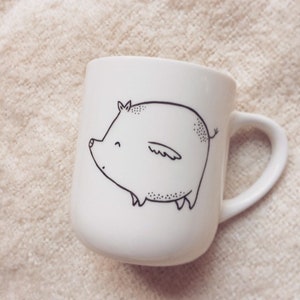 Könnte beinhalten: Weiße Keramiktasse mit einer schwarzen Linienzeichnung eines Schweins mit Flügeln. Das Schwein ist nach links gerichtet und hat einen fröhlichen Ausdruck.