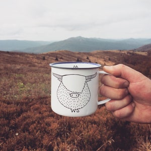 May include: White enamel mug with a black and white illustration of a Highland cow with the text "WS" at the bottom. The mug is being held by a hand against a backdrop of a mountain range.