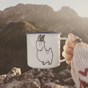 Puede incluir: Taza de camping de esmalte blanco con borde y asa azules. La taza presenta una ilustración negra de una cabra con sombrero. El fondo muestra una cordillera.