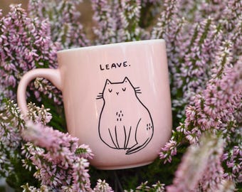 Puede incluir: Una taza de cerámica rosa con una ilustración de gato negro y la palabra "LEAVE". La taza tiene un asa y está rodeada de flores rosas y verdes. La ilustración del gato es sencilla y de estilo caricaturesco.