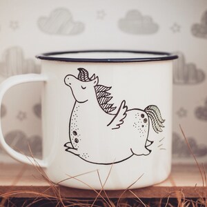 May include: White enamel mug with a black and white illustration of a cartoon unicorn with wings. The mug is sitting in a wooden box with straw.