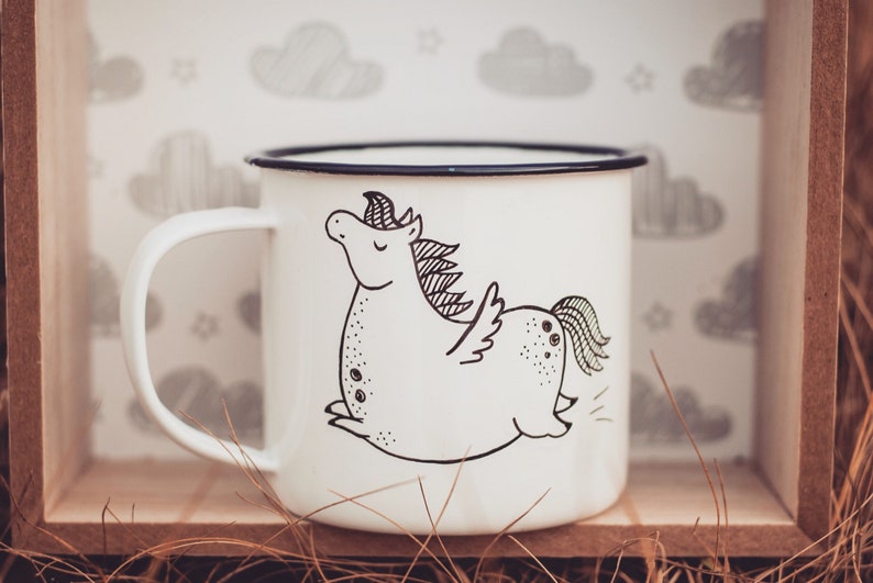 May include: White enamel mug with a black and white illustration of a cartoon unicorn with wings. The mug is sitting in a wooden box with straw.