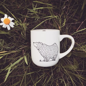 Könnte beinhalten: Weiße Keramiktasse mit einer schwarzen Linienzeichnung eines Bären auf grünem Gras.