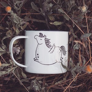 May include: White enamel mug with a black and white illustration of a winged unicorn. The mug is sitting on a bed of dried grasses and flowers.